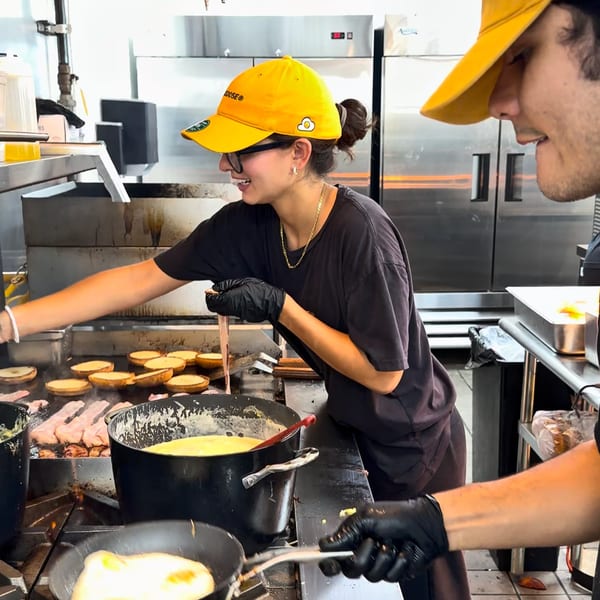 Two chefs in yellow hats and black gloves cook breakfast on a griddle in a busy kitchen.