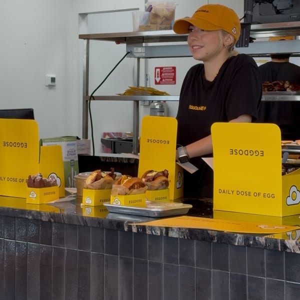 A smiling Eggdose employee in a yellow cap and black shirt stands behind a counter with egg sandwiches.