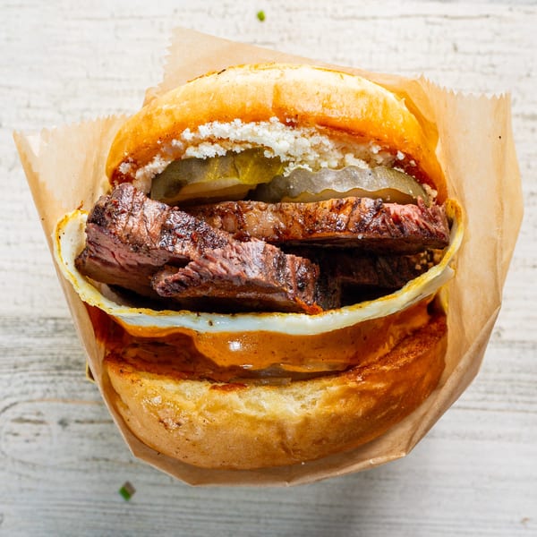 EGGDOSE breakfast sandwich with steak, egg, pickles, and cheese on a bun, wrapped in paper.
