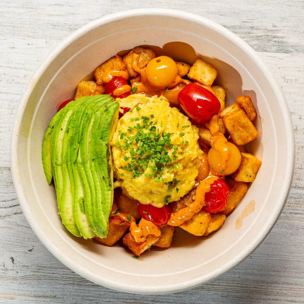 EGGDOSE breakfast bowl with scrambled eggs, avocado, tomatoes, and crispy potatoes in a paper bowl.