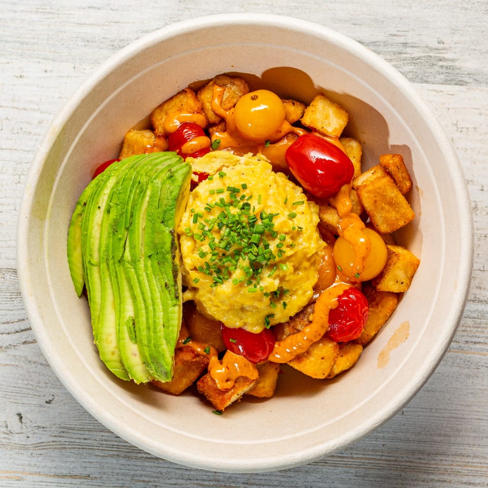 EGGDOSE breakfast bowl with scrambled eggs, avocado, tomatoes, and crispy potatoes in a paper bowl.