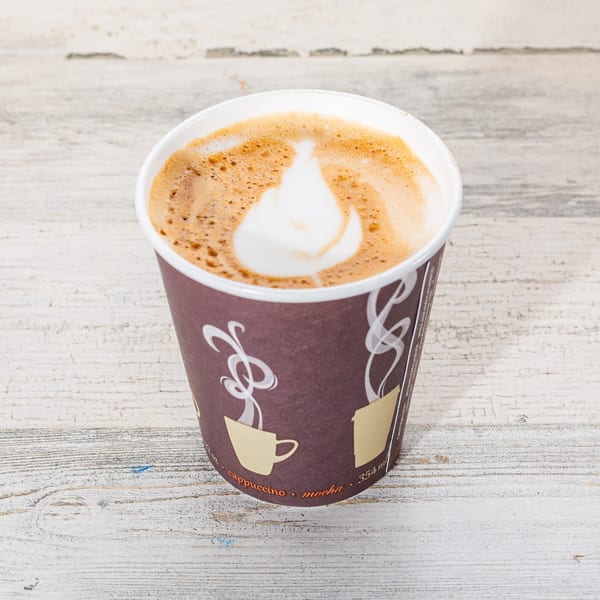 A takeaway cup of cappuccino with latte art on a distressed white wooden surface.