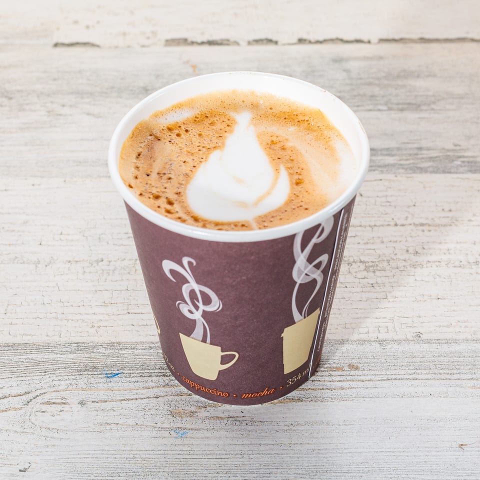 A takeaway cup of cappuccino with latte art on a distressed white wooden surface.