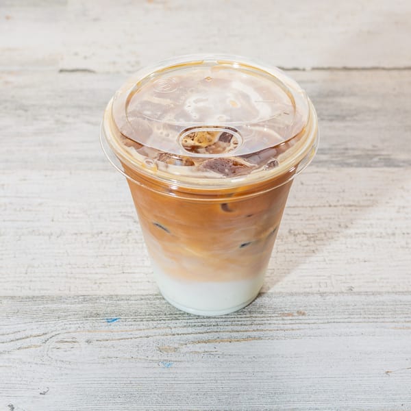 An iced coffee with milk and ice in a clear plastic cup with a lid on a white wooden surface.